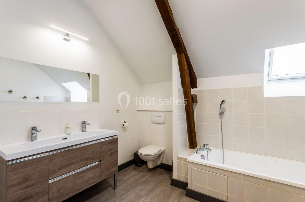 Salle de bain lumineuse avec baignoire, meuble double vasque en bois, toilettes et poutres apparentes.