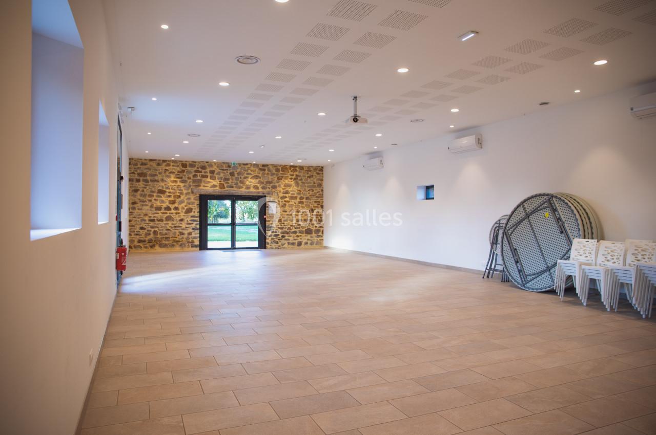Salle vide avec murs blancs, mur en pierre au fond, sol carrelé, chaises empilées et tables pliées sur le côté.