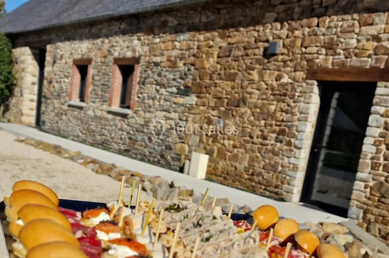 Plateau de bouchées apéritives variées devant un bâtiment en pierre avec toit en ardoise, sous un ciel bleu.