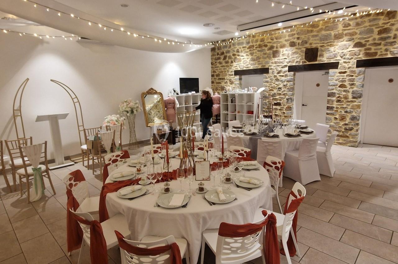 Salle de réception décorée avec des tables rondes, nappes blanches, chaises ornées de rubans rouges et guirlandes lumineuses.