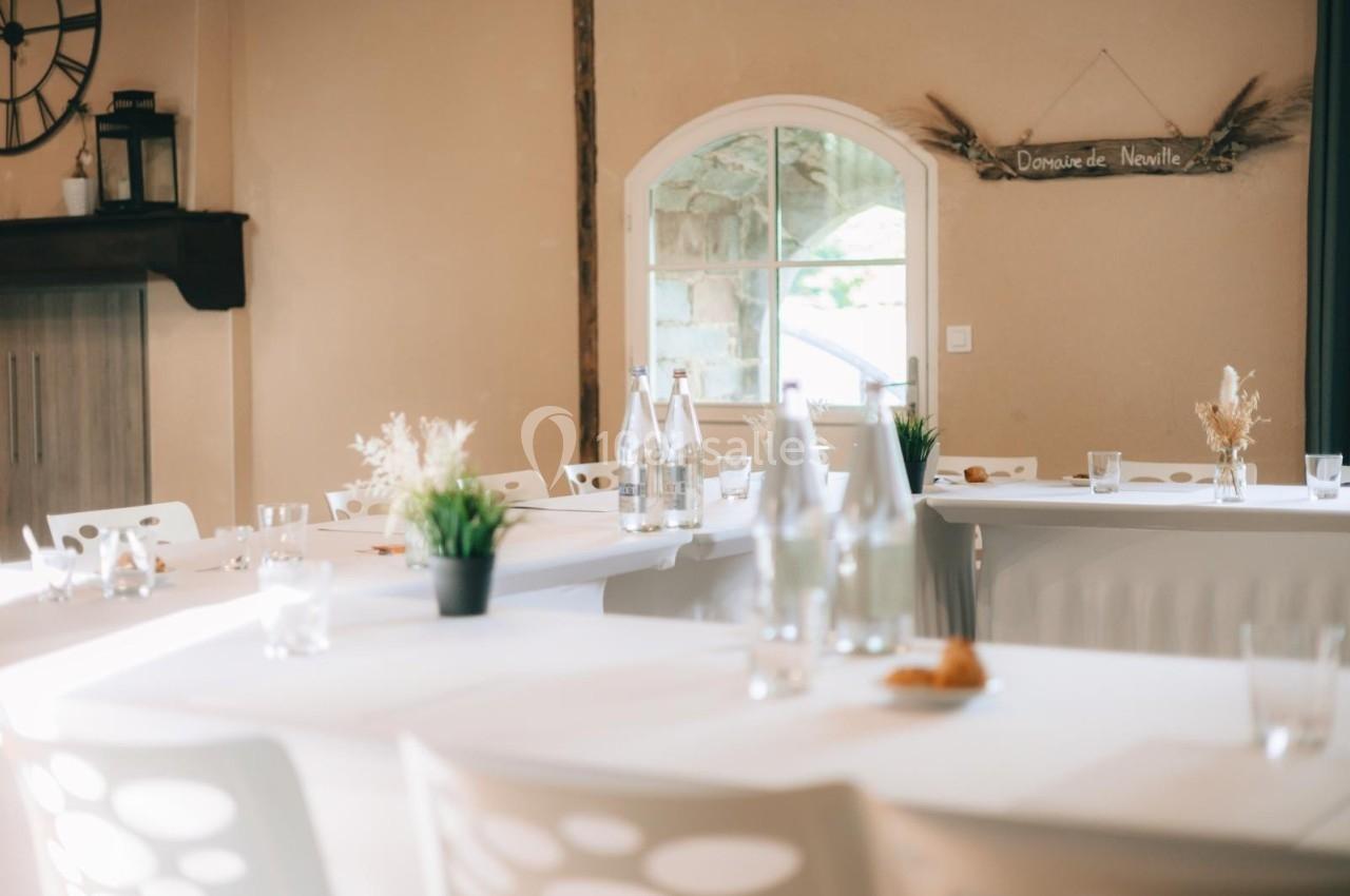 Salle lumineuse avec tables blanches décorées de plantes et bouteilles, près d'une fenêtre et d'un panneau mural.