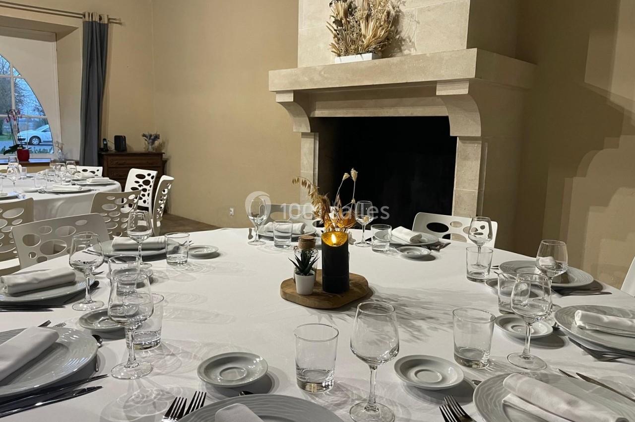 Table dressée avec nappes blanches, vaisselle élégante et centre de table décoratif, dans une salle avec cheminée en pierre.