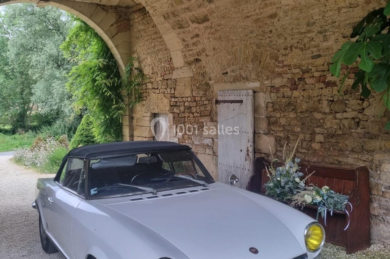Voiture ancienne blanche décapotable garée sous une arche en pierre, entourée de végétation et de décorations florales.