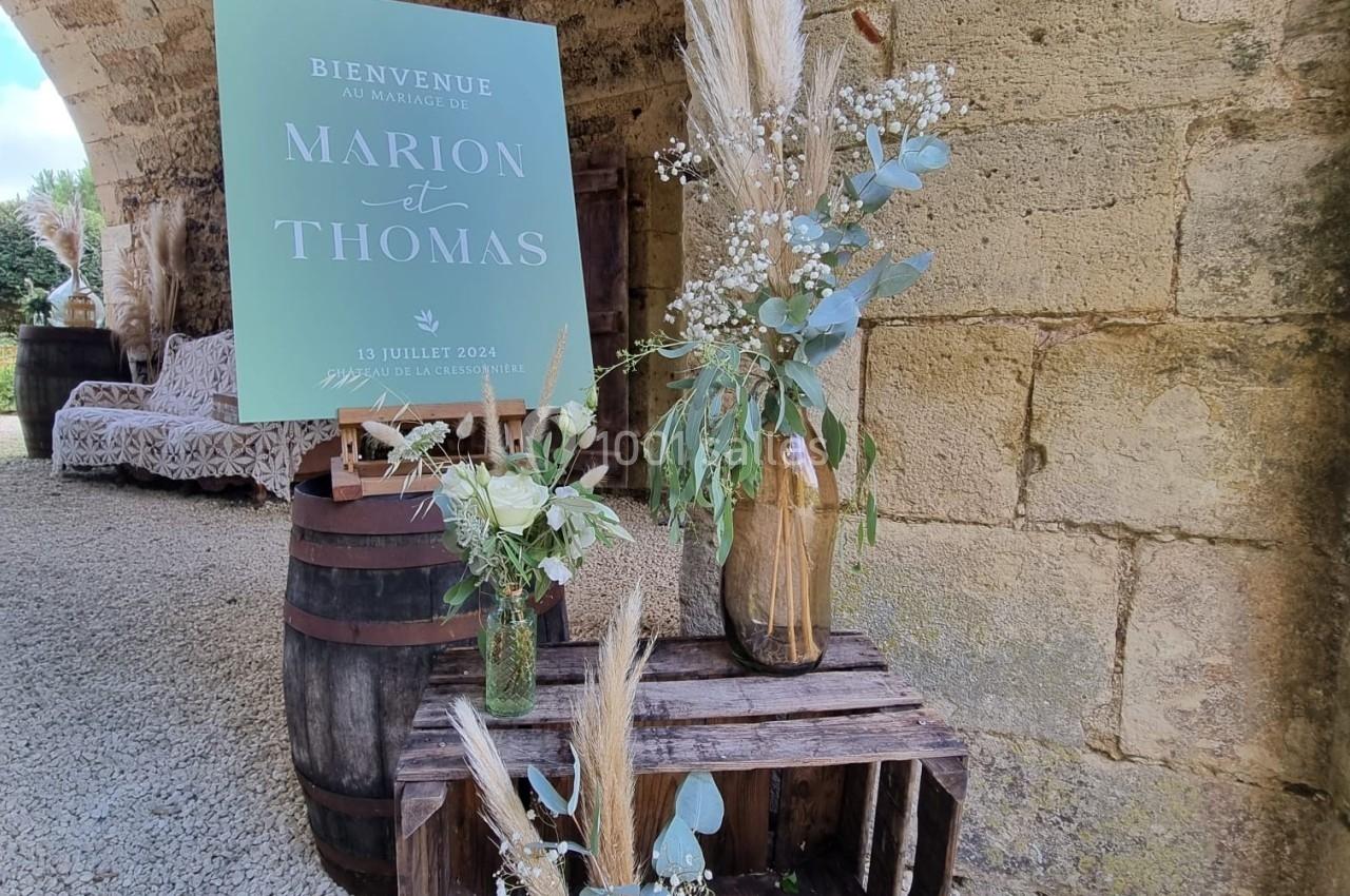 Décoration de mariage avec fleurs séchées, panneaux en bois, et affiche personnalisée sous une arche en pierre.