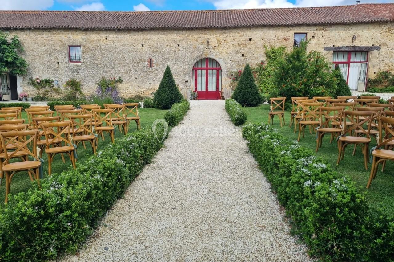 Allée centrale bordée de buissons menant à une porte rouge, entourée de chaises en bois dans un jardin.