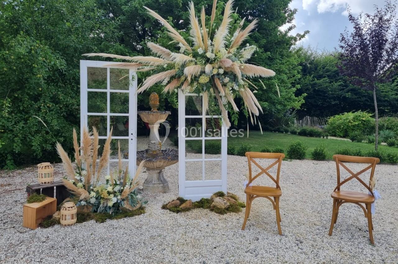 Décor extérieur avec portes blanches, fleurs séchées, fontaine centrale et deux chaises en bois sur gravier.