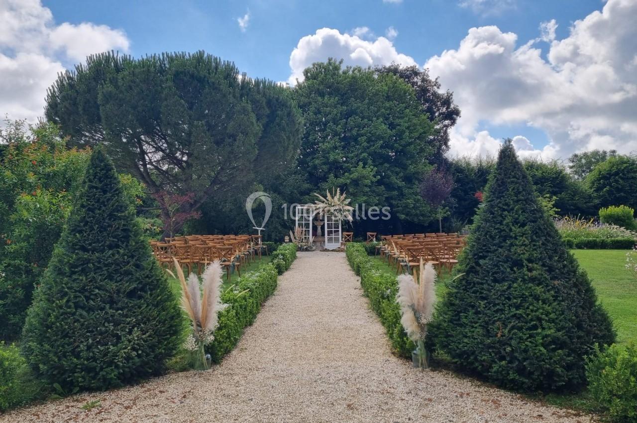 Allée centrale bordée de verdure menant à une arche décorée pour une cérémonie en plein air, sous un ciel partiellement…