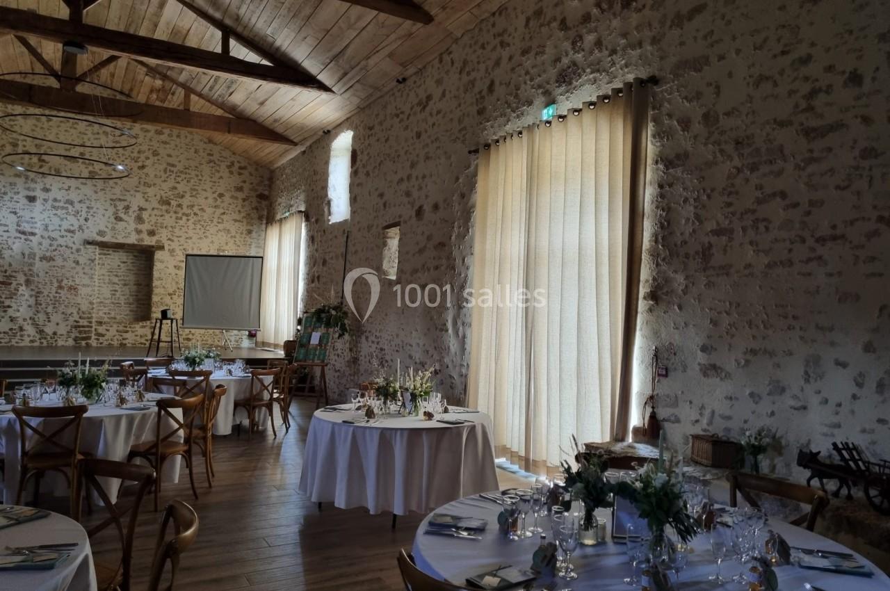 Salle de réception en pierre avec tables rondes dressées, nappes blanches et lumière naturelle filtrée par des rideaux…