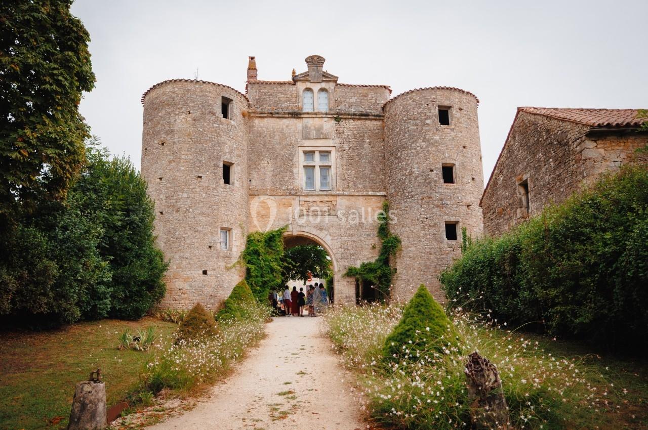 Entrée d'un château en pierre avec deux tours rondes, entouré de verdure et d'un chemin bordé de fleurs.