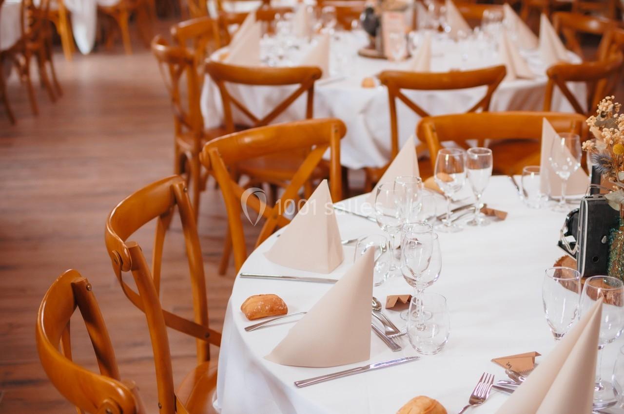 Salle de réception avec tables rondes dressées, nappes blanches, serviettes pliées et chaises en bois.