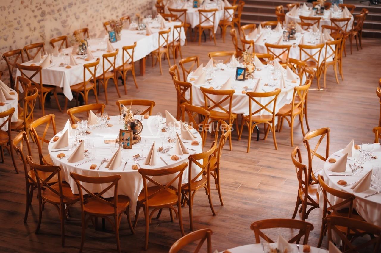 Salle de réception avec des tables rondes dressées, nappes blanches, chaises en bois et décorations florales.