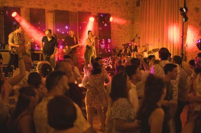 Un homme debout, bras ouverts, s'exprime devant un mur en pierre, avec une chaise en bois à côté de lui.
