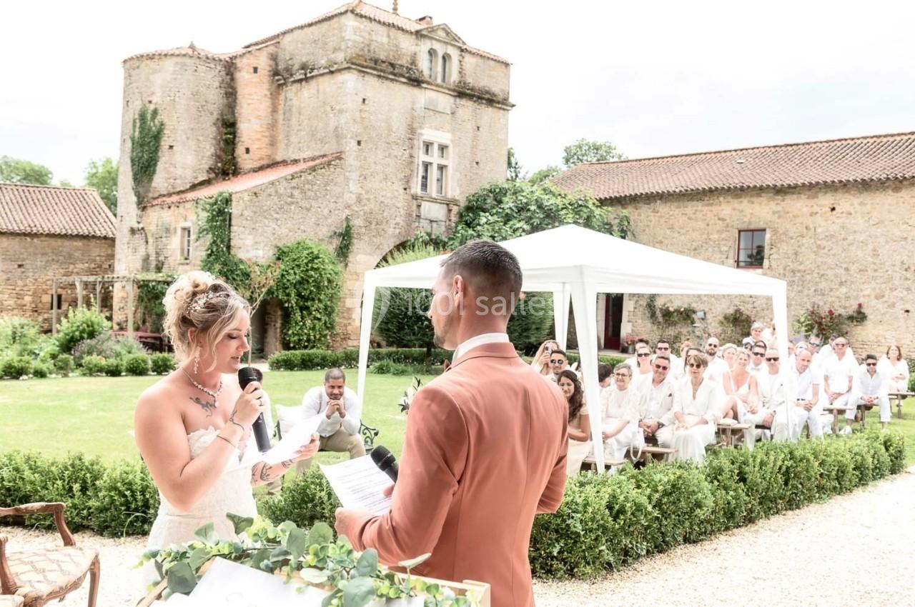 Une mariée lit un discours au micro face à son époux lors d'une cérémonie en plein air devant des invités.