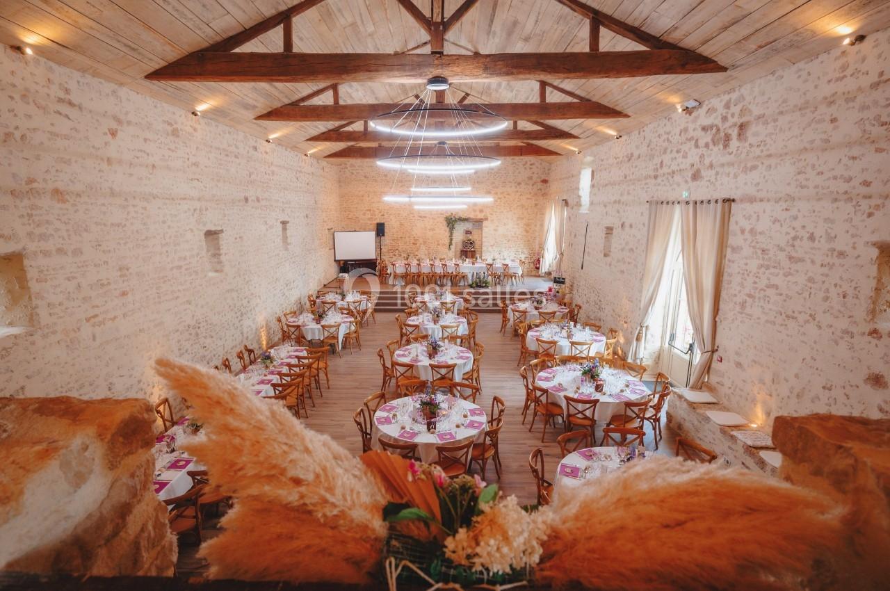 Salle de réception en pierre avec tables rondes décorées, chaises en bois et plafonds avec poutres apparentes.