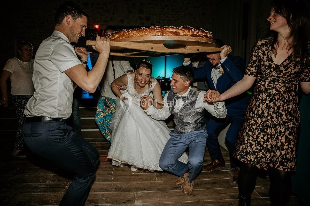 Des mariés souriants sont soulevés sous une grande brioche portée par des invités dans une ambiance festive.