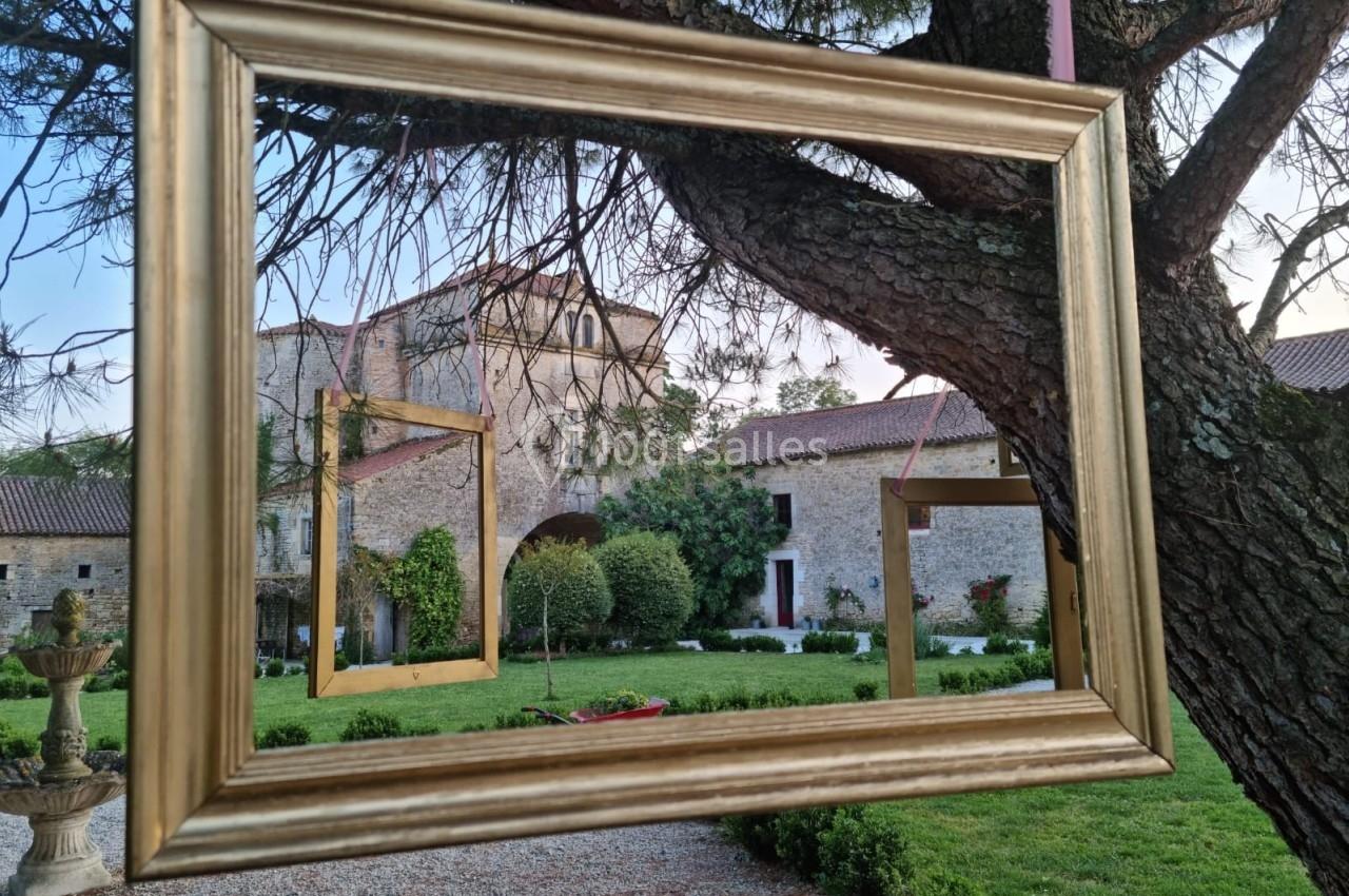 Cadres dorés suspendus à un arbre, encadrant une vue sur un jardin et un bâtiment en pierre.