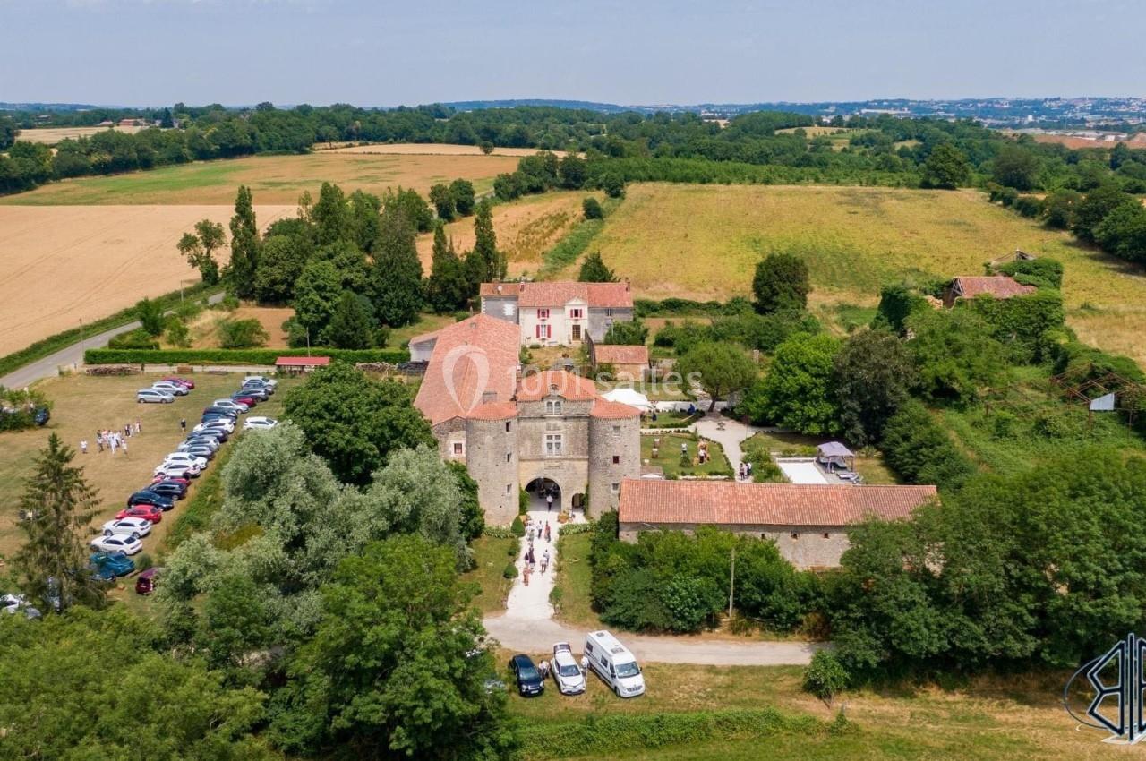 Vue aérienne d'un domaine rural avec bâtiments en pierre, entouré de champs, de voitures stationnées et de végétation.