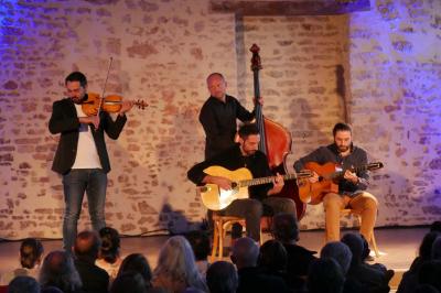 Un homme debout, bras ouverts, s'exprime devant un mur en pierre, avec une chaise en bois à côté de lui.