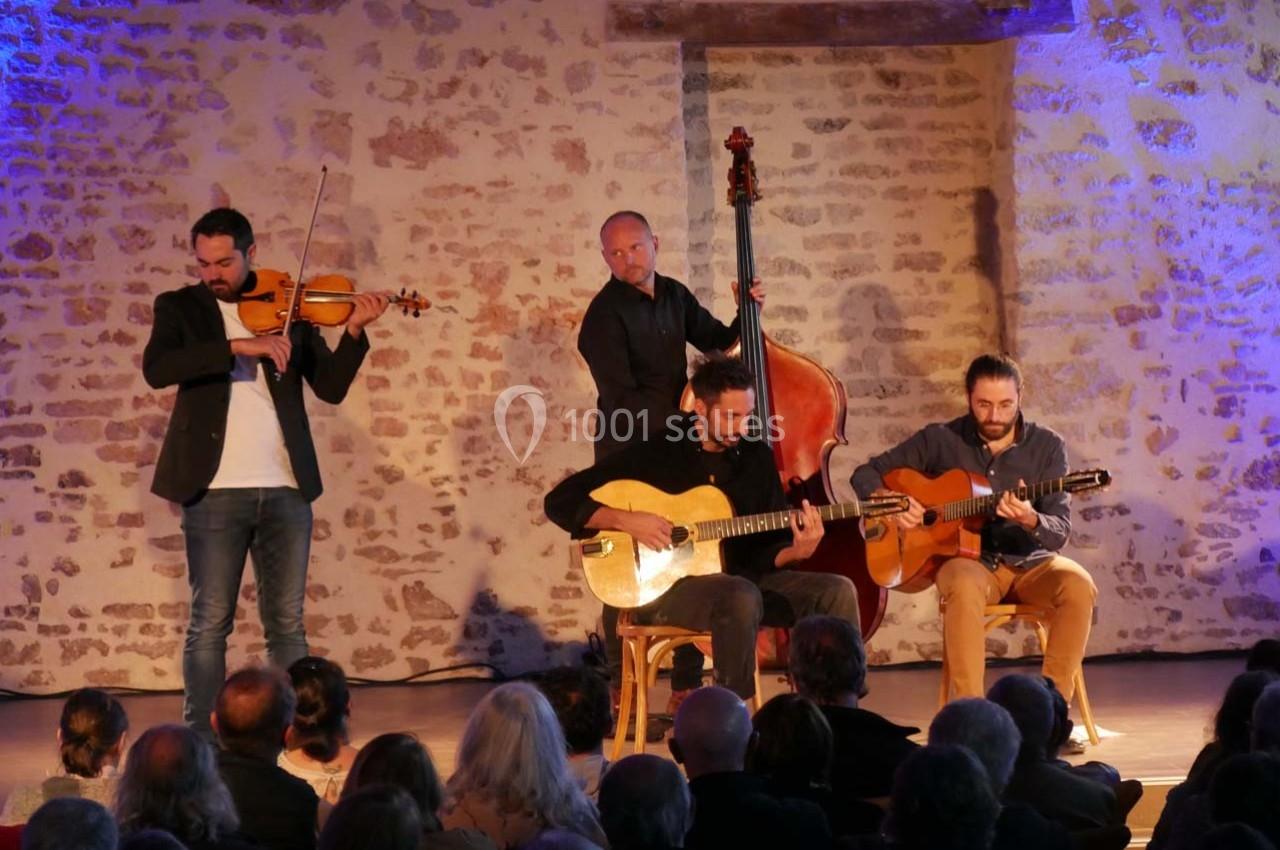 Quatuor de musiciens jouant du violon, de la contrebasse et des guitares devant un public dans une salle en pierre.