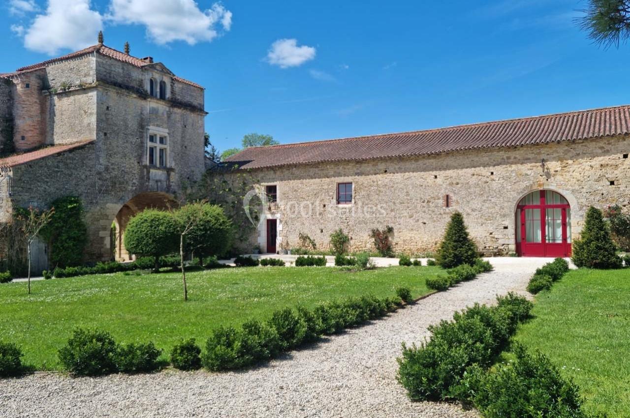 Bâtiment en pierre avec toit en tuiles, jardin aménagé et allée gravillonnée sous un ciel bleu.