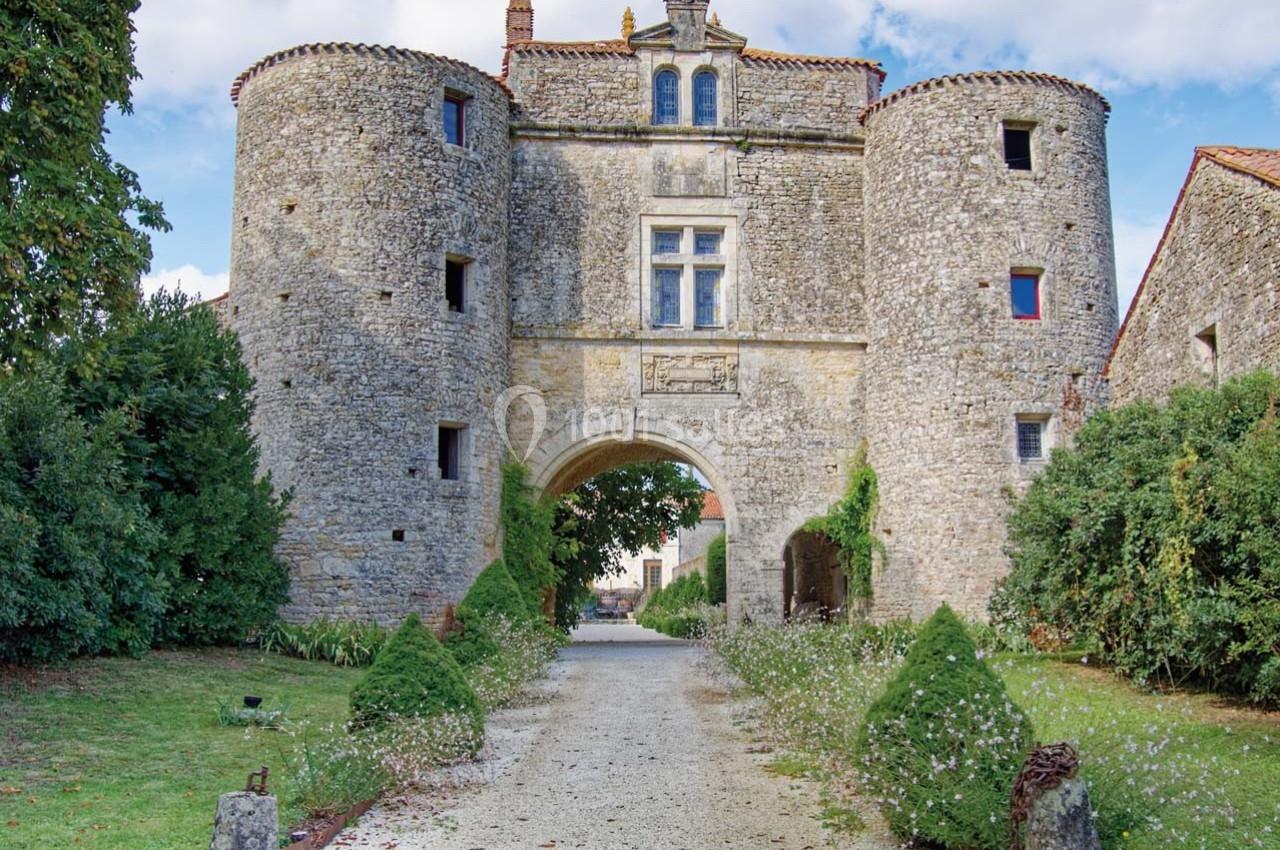 Château en pierre avec deux tours rondes, une arche centrale et un chemin bordé de verdure.