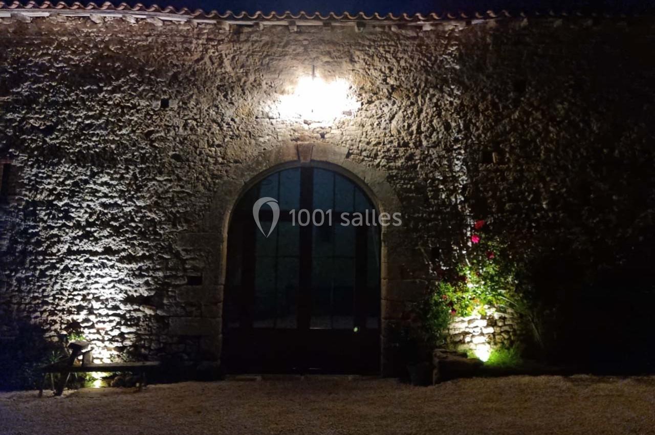 Façade en pierre éclairée de nuit avec une grande porte vitrée en arc et des plantes grimpantes sur le côté.