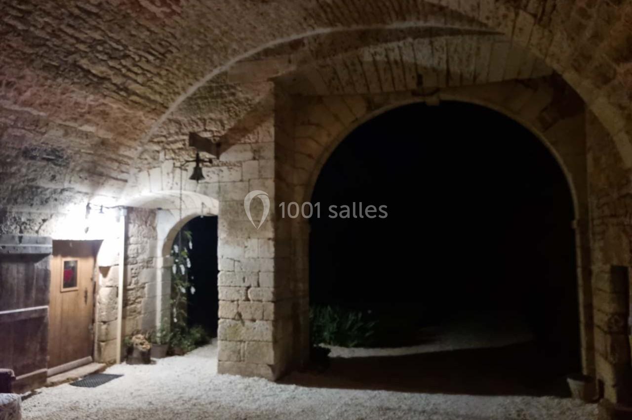 Entrée voûtée en pierre éclairée, avec une porte en bois à gauche et une ouverture sombre donnant sur l'extérieur.