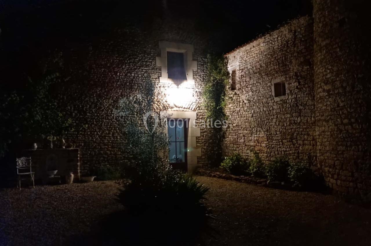 Façade en pierre éclairée la nuit, avec une porte vitrée centrale et des plantes au premier plan.