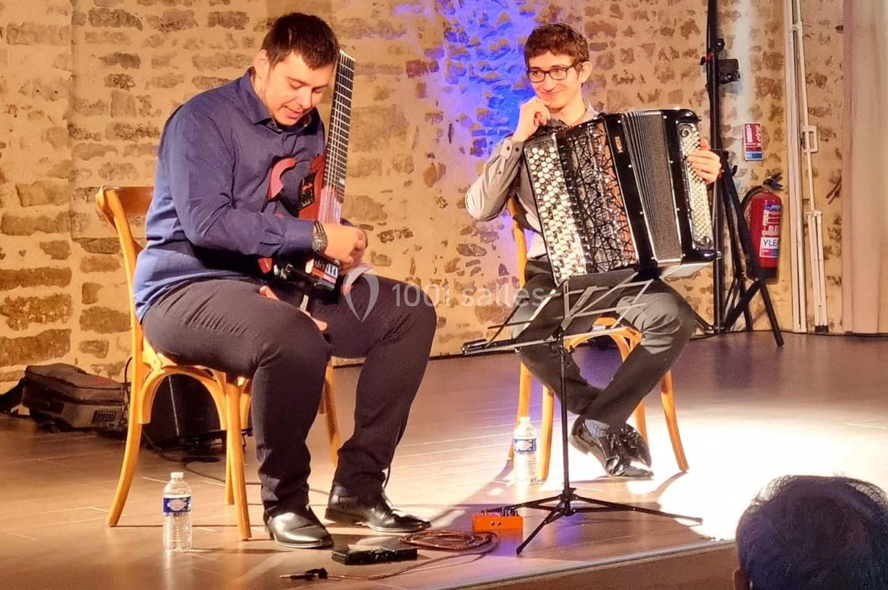 Deux musiciens assis jouent de la guitare électrique et de l'accordéon sur une scène éclairée dans une salle en pierre.
