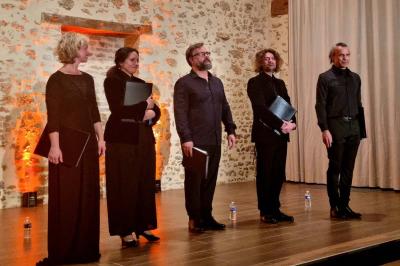 Un homme debout, bras ouverts, s'exprime devant un mur en pierre, avec une chaise en bois à côté de lui.
