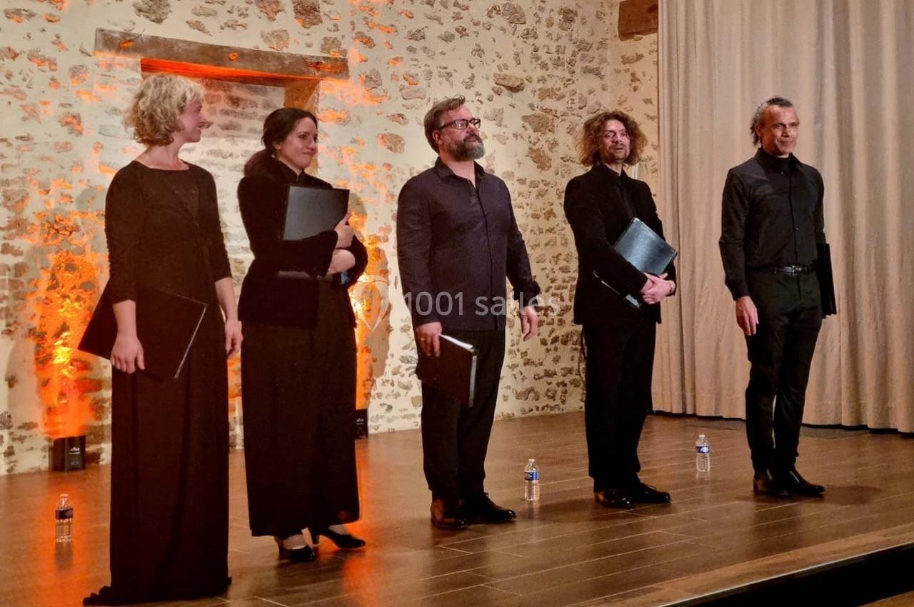 Cinq chanteurs en tenue de concert debout sur une scène en bois, devant un mur en pierre éclairé.