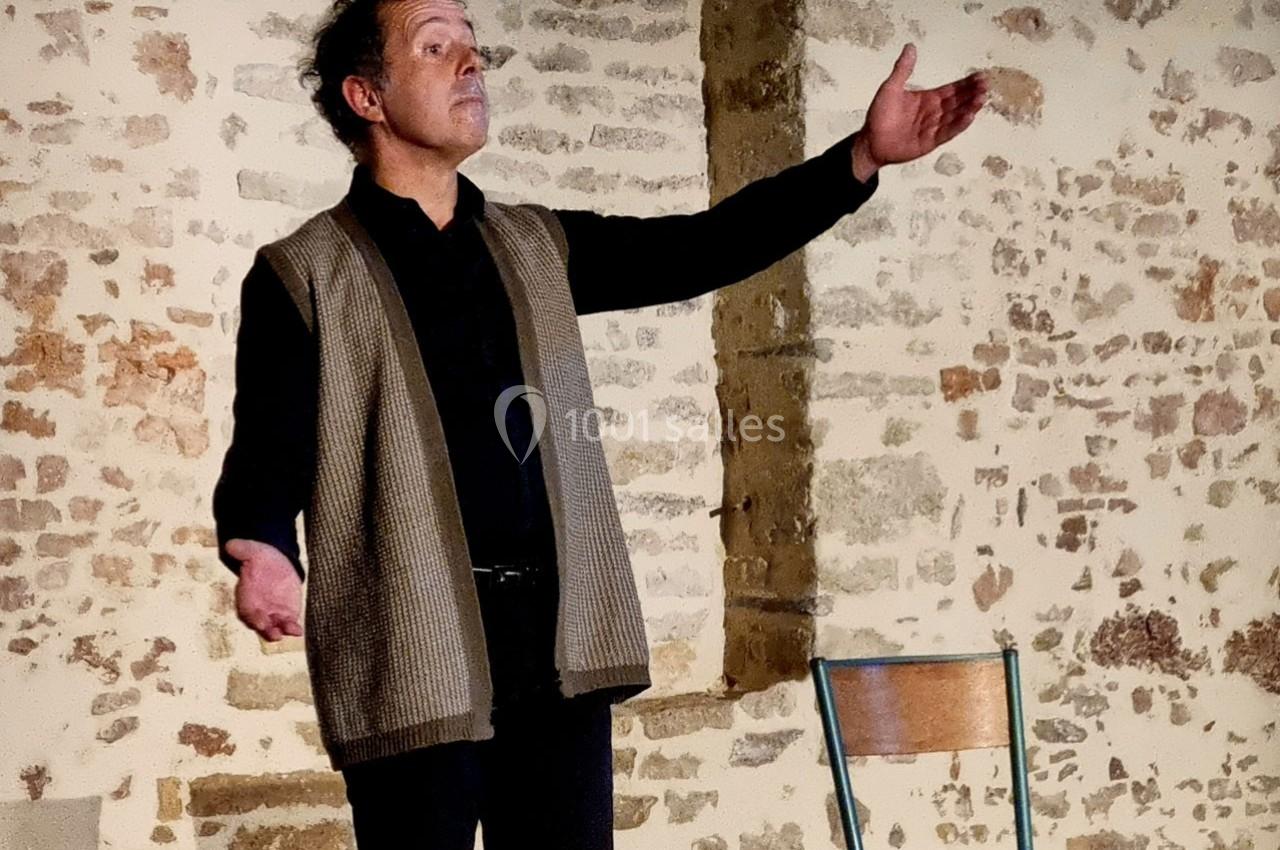 Un homme debout, bras ouverts, s'exprime devant un mur en pierre, avec une chaise en bois à côté de lui.