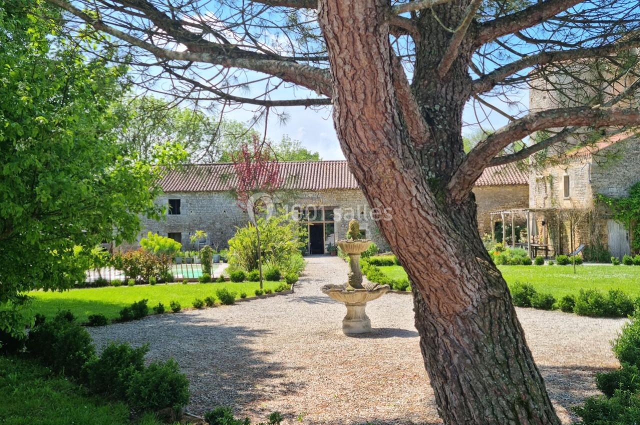 Cour d'une maison en pierre avec fontaine centrale, jardin verdoyant et arbres sous un ciel ensoleillé.
