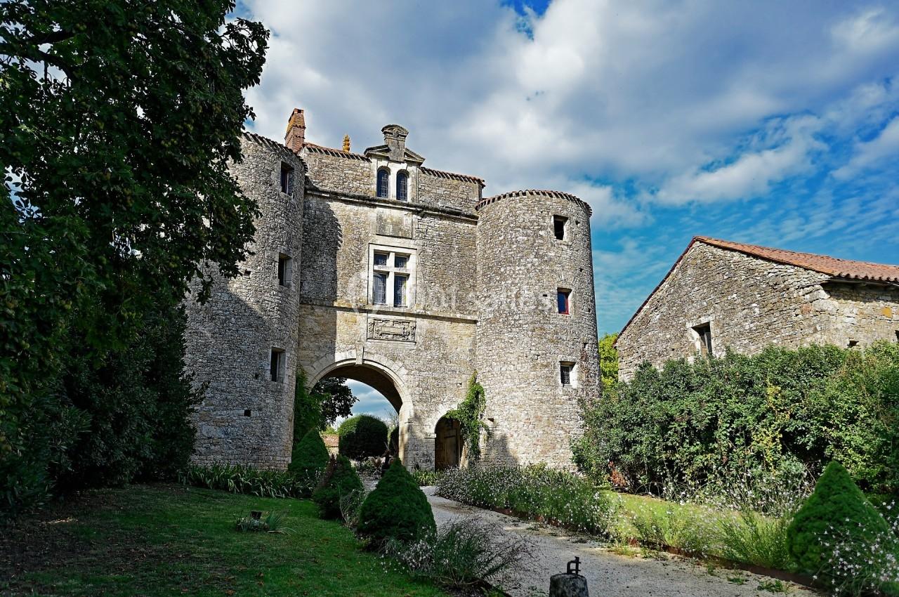 Entrée d'un château en pierre avec deux tours rondes, entouré de verdure sous un ciel partiellement nuageux.