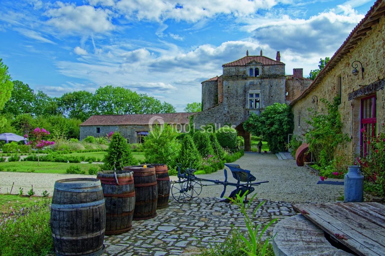 Cour pavée avec tonneaux, charrette et jardin fleuri, menant à un bâtiment en pierre sous un ciel partiellement nuageux.