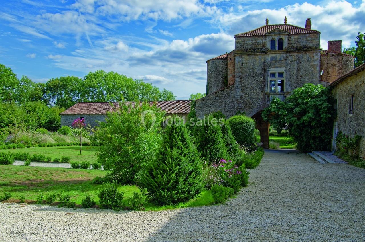 Allée gravillonnée menant à un bâtiment en pierre entouré de jardins verdoyants sous un ciel partiellement nuageux.