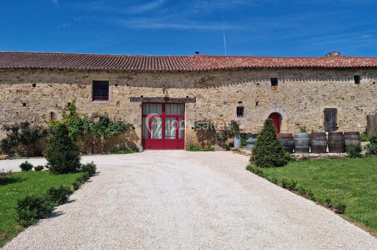 Façade en pierre d'un bâtiment ancien avec porte rouge, barriques en bois et jardin aménagé sous un ciel bleu.
