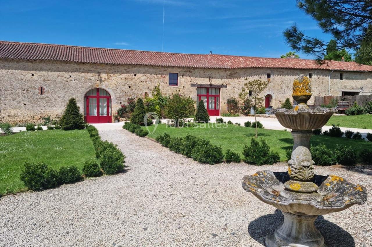 Cour d'une bâtisse en pierre avec fontaine au premier plan, allées gravillonnées et jardin paysager.