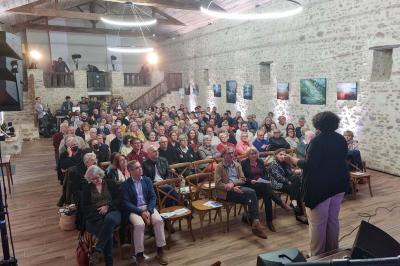 Un homme debout, bras ouverts, s'exprime devant un mur en pierre, une chaise vide à ses côtés.