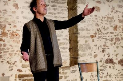 Un homme debout, bras ouverts, s'exprime devant un mur en pierre, une chaise vide à ses côtés.