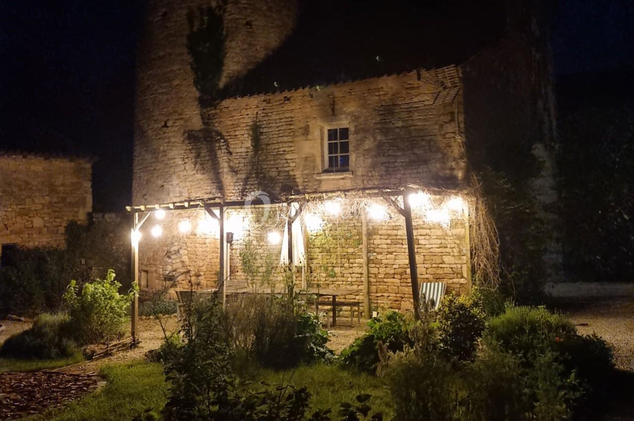 Pergola éclairée par des guirlandes lumineuses devant une maison en pierre, entourée de végétation, de nuit.