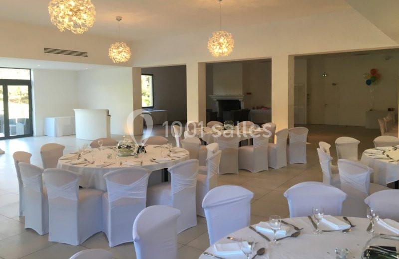 Salle de réception lumineuse avec des tables rondes dressées, entourées de chaises blanches houssées.