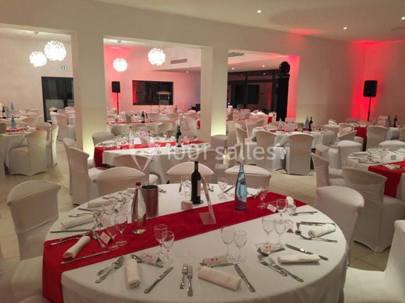 Salle de réception décorée avec des tables rondes, nappes blanches, chemins de table rouges et chaises habillées.