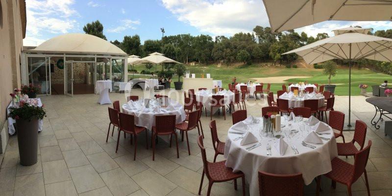Terrasse aménagée avec des tables rondes dressées pour un événement, entourée de parasols et donnant sur un terrain…