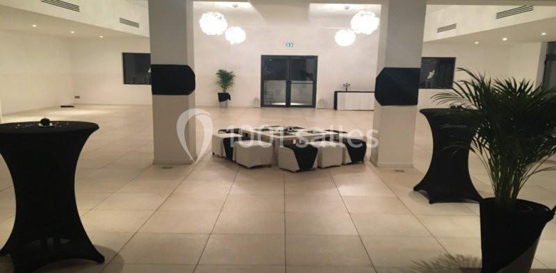 Salle spacieuse avec sol carrelé, tables hautes noires, poufs blancs et noirs, et plantes décoratives.