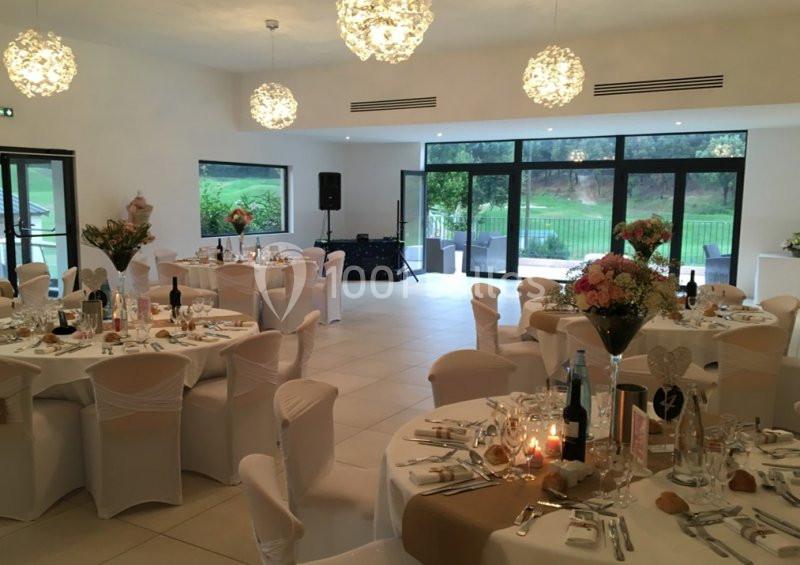 Salle de réception élégamment décorée avec des tables rondes dressées, des chaises blanches et une vue sur un jardin.