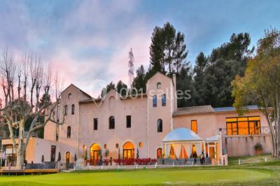 Bâtiment en pierre de style méditerranéen entouré d'arbres, avec une terrasse et une pelouse au premier plan.