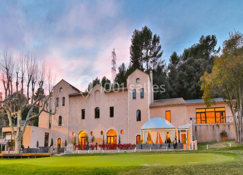 Bâtiment en pierre de style méditerranéen entouré d'arbres, avec une terrasse et une pelouse au premier plan.