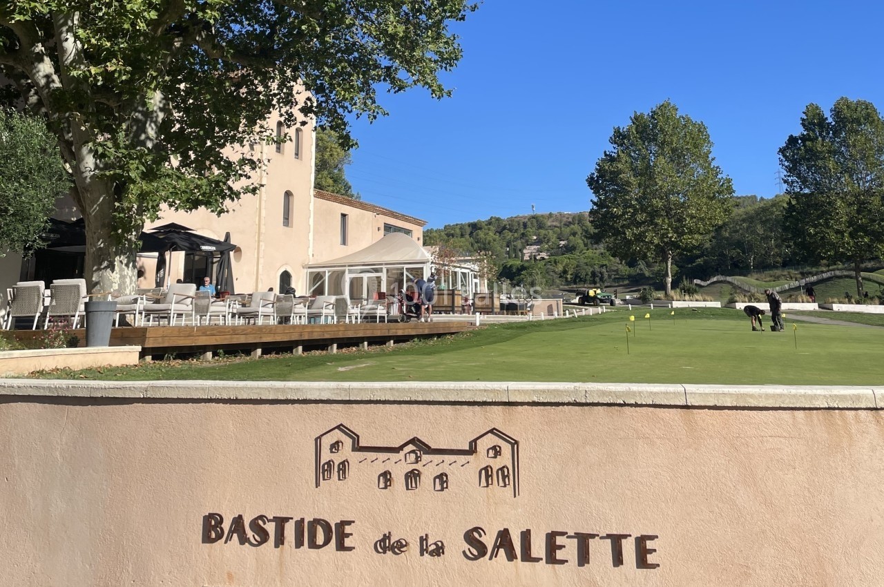 Location salle Marseille 11 (Bouches-du-Rhône) - La Bastide de la Salette #12 Terrasse d'un bâtiment beige avec des tables, vue sur un terrain de golf et un panneau ’Bastide de la Salette’.