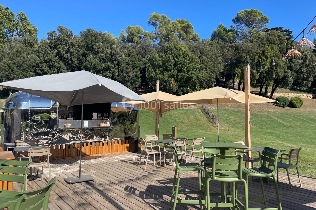 Location salle Marseille 11 (Bouches-du-Rhône) - La Bastide de la Salette #8 Terrasse en bois avec tables, chaises et parasols, située près d'un food truck métallique dans un cadre verdoyant.